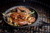 Solidteknics Iron Crêpe Pan cooking steak and onions on a Big Green Egg grill, showcasing exceptional heat retention and versatility.